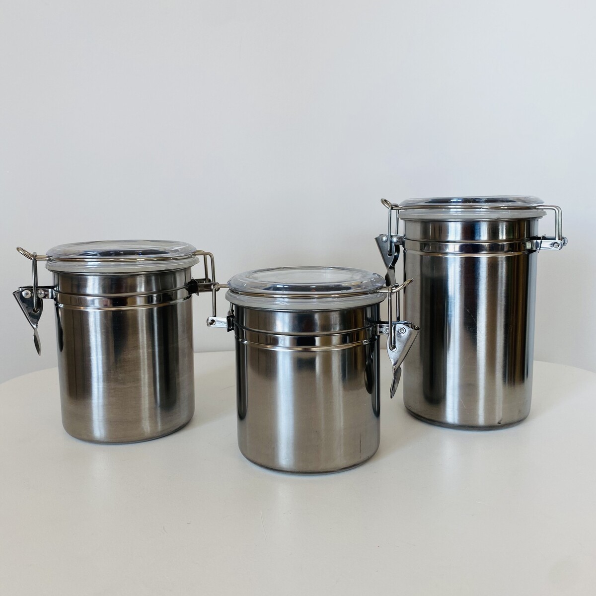 Três recipientes cilíndricos de aço inox com tampas transparentes e fechos metálicos