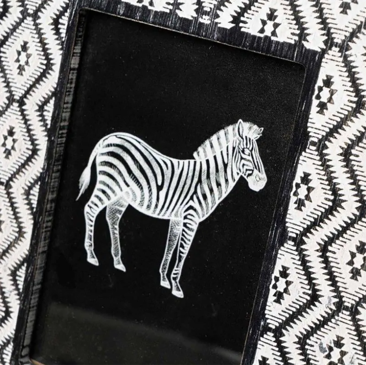 Placa decorativa com imagem de zebra em fundo preto e moldura preta sobre padrão geométrico branco e preto