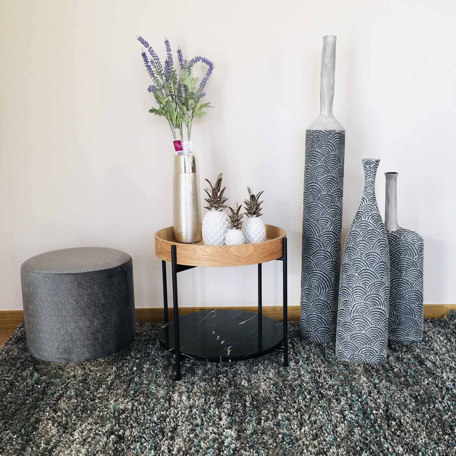 Mesa de apoio redonda com tampo de madeira e vidro preto, três jarrões cinzentos altos com padrões, puff cinzento e decoração sobre tapete cinzento.