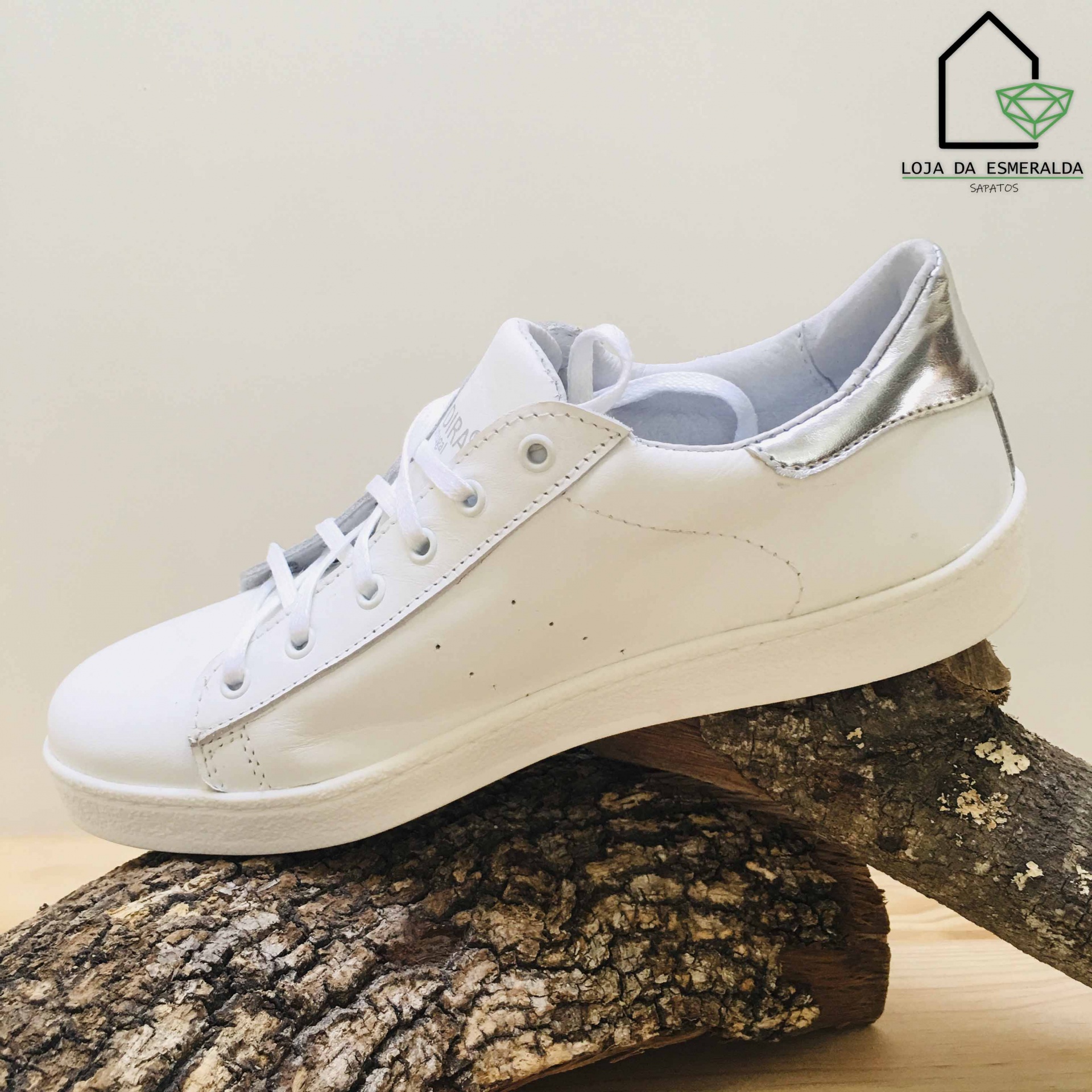 Ténis branco com detalhes prateados sobre tronco de árvore com logótipo Loja da Esmeralda