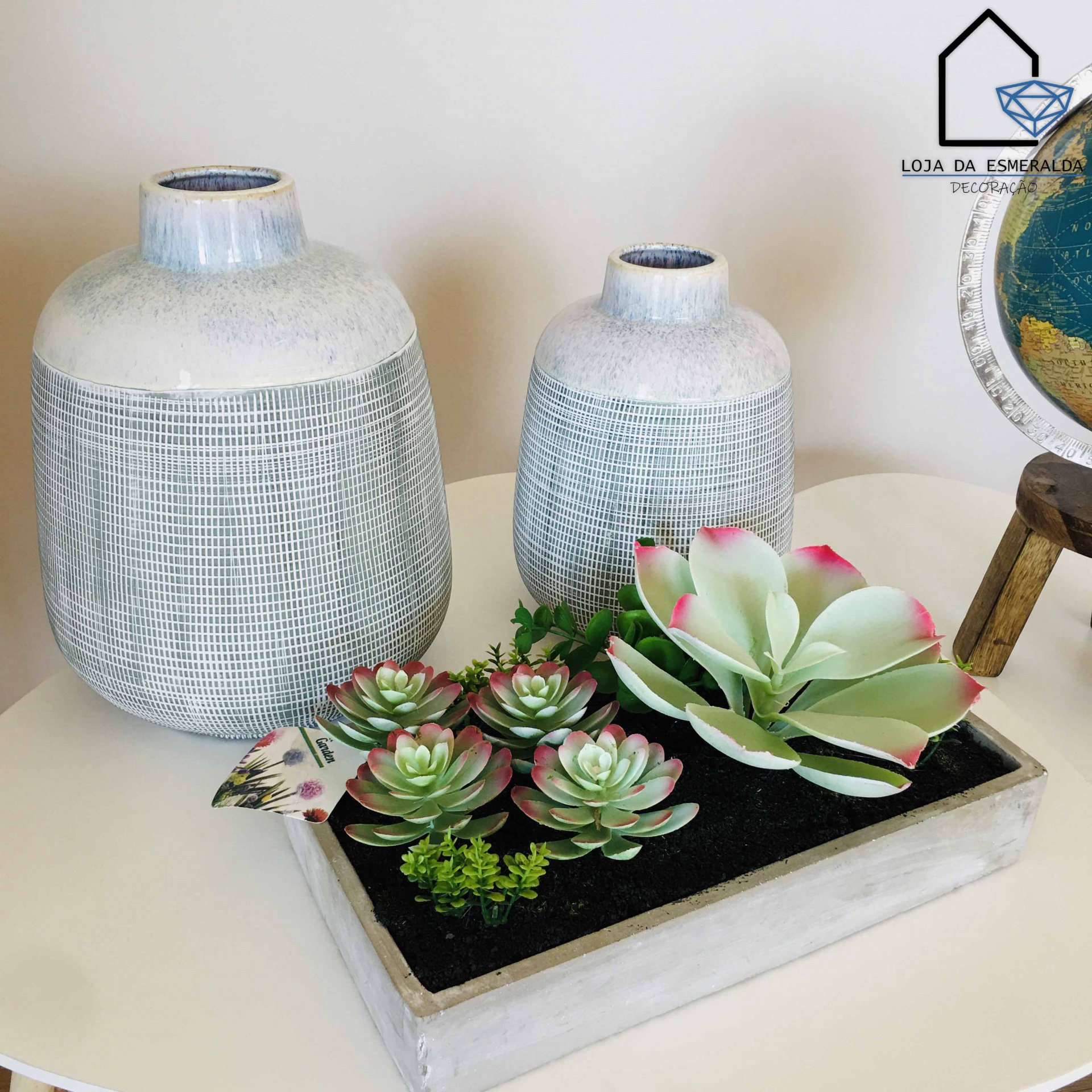Dois vasos decorativos em tons cinza e branco com padrão de linhas, bandeja com suculentas artificiais verdes e rosa sobre mesa branca.