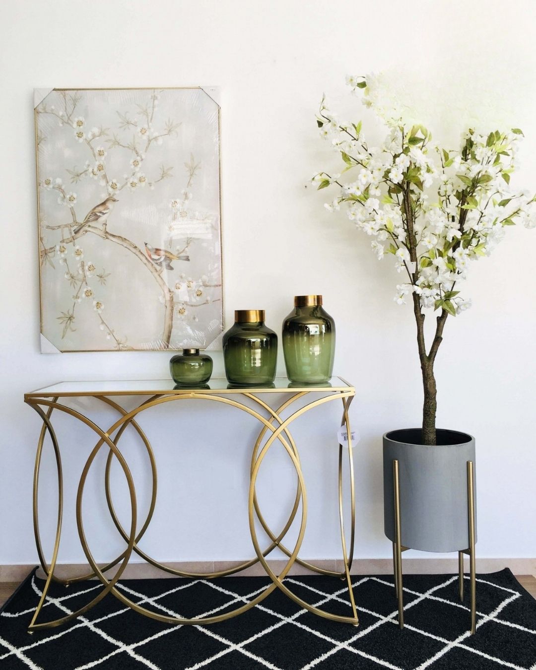 Mesa de entrada dourada com vidro e decoração com jarras verdes, quadro de pássaros e planta artificial branca
