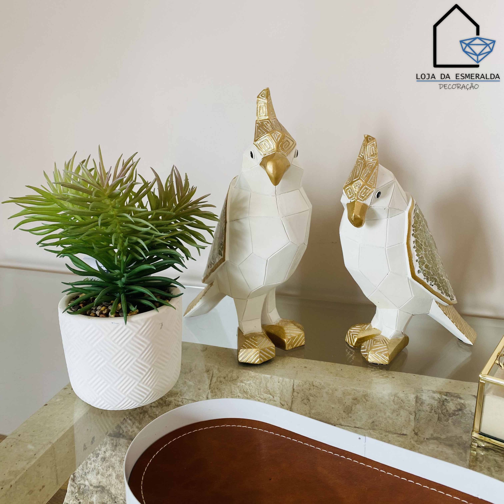 Dois ornamentos de papagaios branco e dourado ao lado de planta em vaso branco sobre mesa com superfície de pedra