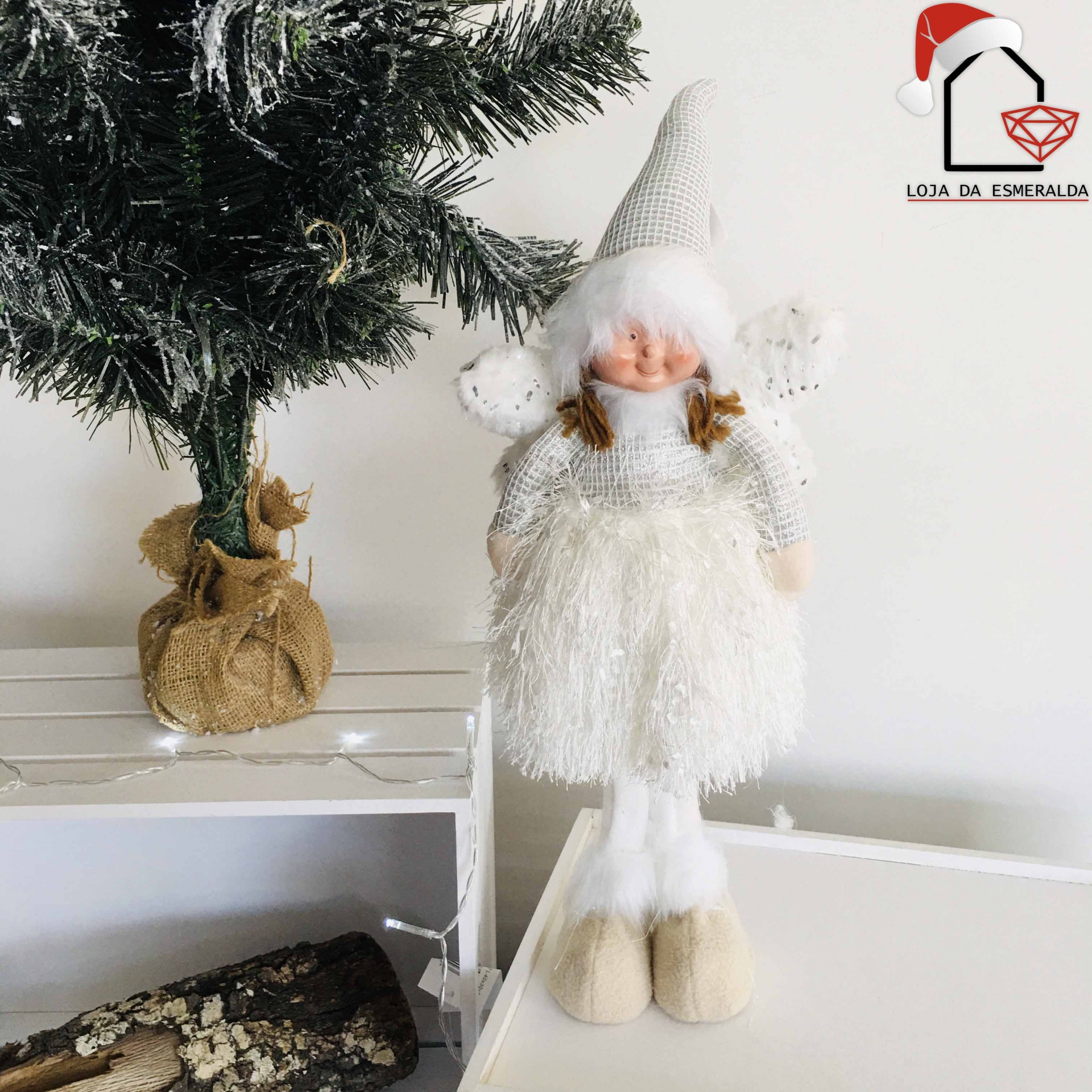 Boneco decorativo de Natal com roupa branca felpuda e chapéu pontudo cinzento ao lado de árvore de Natal artificial.