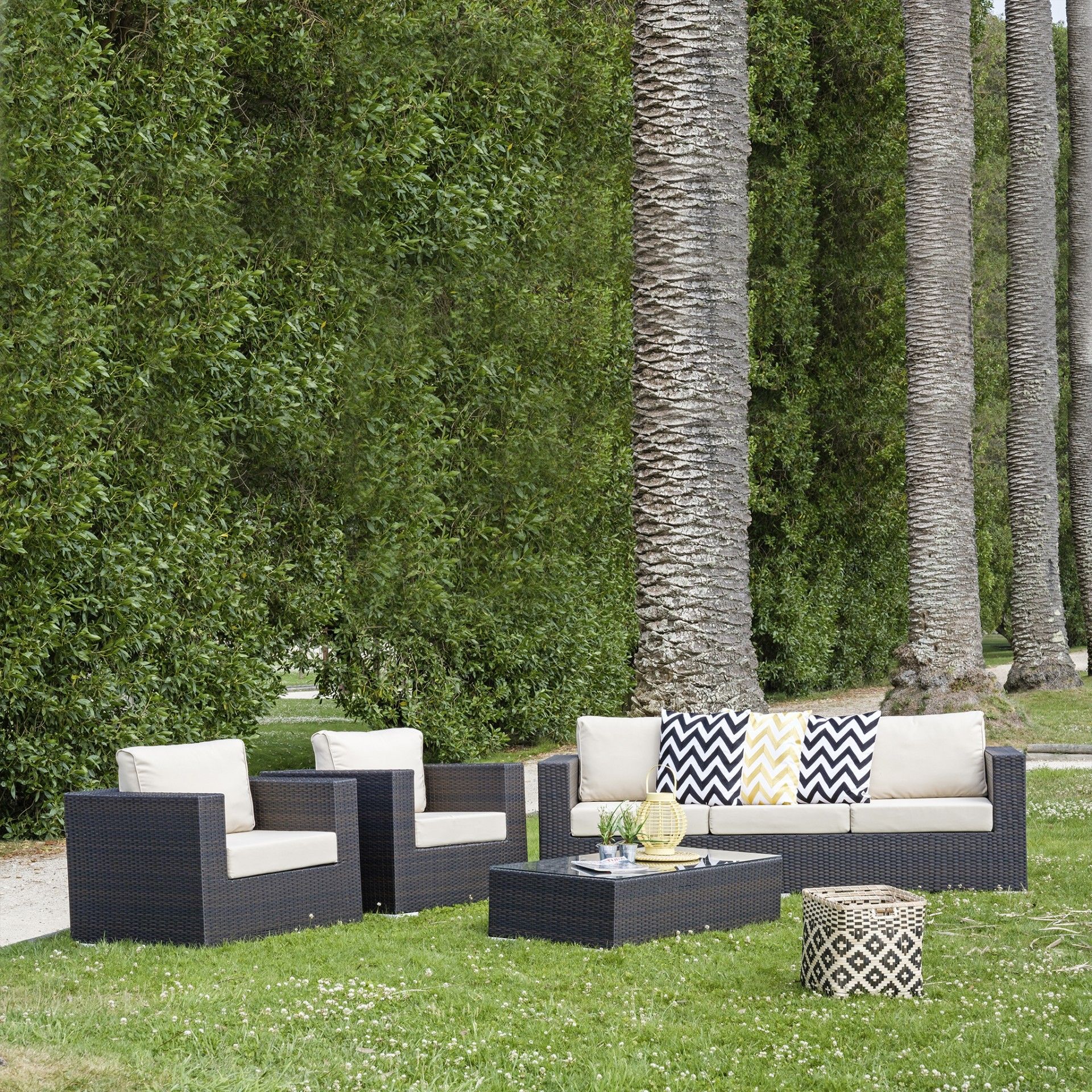 Conjunto de mobiliário de exterior em rattan sintético preto com almofadas claras e decorativas em relva com palmeiras.