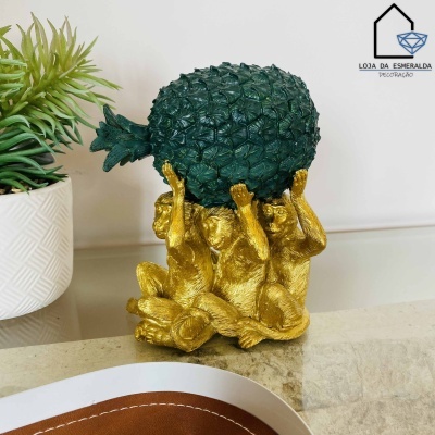 Escultura de três macacos dourados segurando um abacaxi verde sobre superfície clara com planta ao fundo