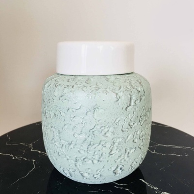 Jarro decorativo verde água com tampa branca sobre superfície preta com padrão branco