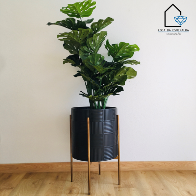 Vaso de planta artificial em suporte metálico numa sala com chão de madeira.