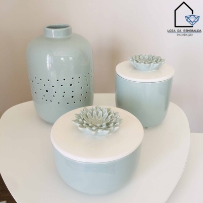 Conjunto de cerâmica verde clara com vaso perfurado e potes com flores decorativas em tampas brancas sobre mesa branca