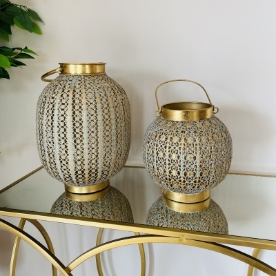 Dois lanternas decorativos de metal dourado e branco com padrão rendado sobre mesa de vidro dourada