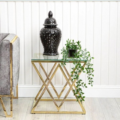 Mesa lateral dourada com tampo de vidro, vaso preto decorativo e planta verde ao lado de sofá cinza