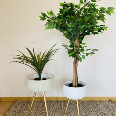 Dois vasos de plantas artificiais em suportes de madeira com plantas diferentes numa sala com parede creme e tapete castanho