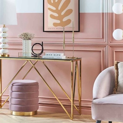 Conjunto de mobiliário com secretária de vidro e dourado, puff rosa e sofá com manta bege em parede rosa