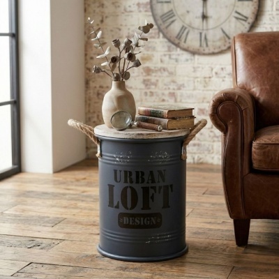 Mesa lateral de tambor metálico cinzento com vaso, lupa e livros ao lado de sofá de couro