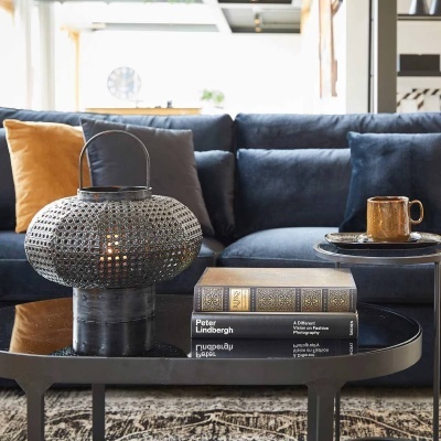 Mesa de centro com lanterna metálica preta, livros empilhados e caneca castanha, sofá azul ao fundo