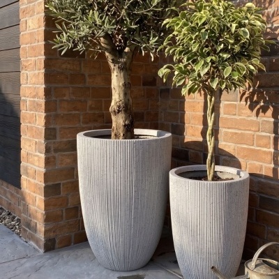 Dois vasos cinza com textura de linhas verticais e árvores plantadas junto a uma parede de tijolos com ferramentas de jardinagem no chão.