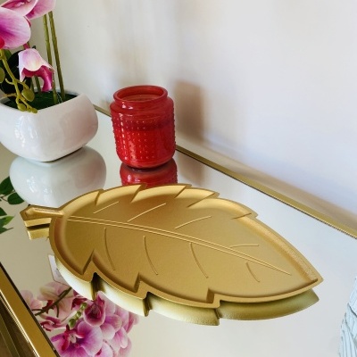 Placa dourada em forma de folha numa superfície espelhada com vaso de flores cor-de-rosa e vela vermelha