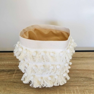 Saco decorativo em tecido branco com franjas e pompons colocado numa superfície de madeira.