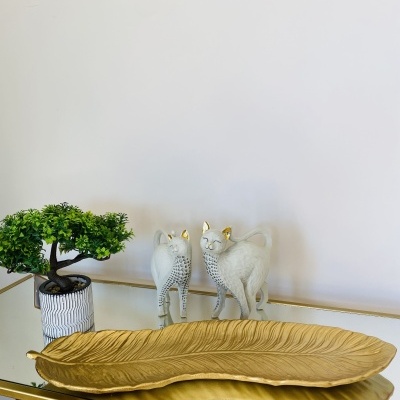 Decoração com placa dourada em forma de folha, vaso com planta artificial e esculturas de gatos cinzentos com detalhes dourados.