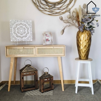 Mesa de madeira clara com gavetas de rattan, lanternas, vaso dourado com flores secas, banco branco e decoração de parede clara