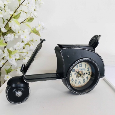 Relógio em formato de scooter vintage preta com mostrador redondo na roda sobre mesa branca próximo a flores brancas