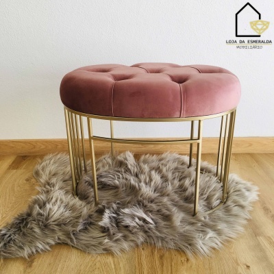 Pufe redondo rosa com base metálica dourada sobre tapete peludo cinza