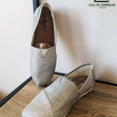 Par de sapatos prateados brilhantes tipo alpargatas na madeira ao lado do logótipo da loja
