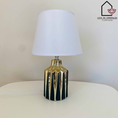 Abajur com base dourada facetada e cúpula branca sobre mesa branca
