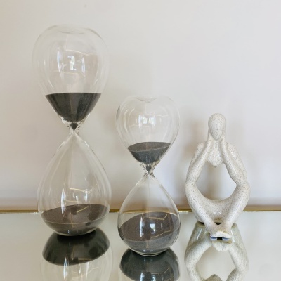 Dois ampulhetas de vidro com areia preta ao lado de uma estátua branca de figura humana sentada.