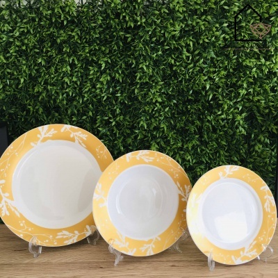 Conjunto de três pratos com bordas amarelas e padrão branco sobre suporte plástico em madeira clara com fundo de plantas verdes