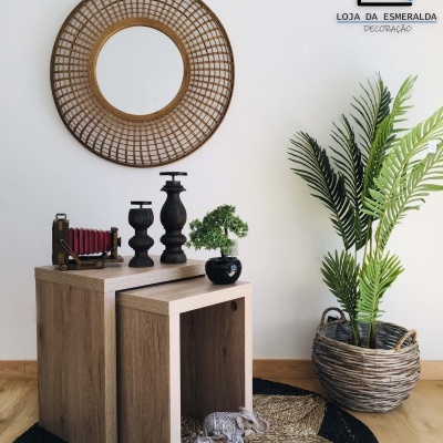 Espelho redondo de rattan, mesas ninho de madeira clara com decoração, plantas artificiais, tapete e logotipo em parede branca.
