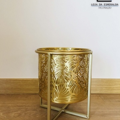Recipiente decorativo metálico dourado com padrão de folhas em suporte dourado sobre chão de madeira
