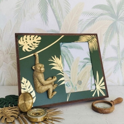 Moldura de foto verde com folhas douradas e macaco decorativo, bússola antiga e lupa de madeira ao lado.
