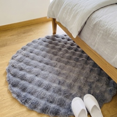 Tapete redondo cinzento felpudo ao lado de cama de madeira clara com roupa de cama branca e chinelos brancos no chão de madeira