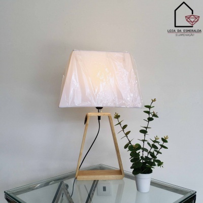 Candeeiro de mesa com cúpula branca e base em madeira clara sobre mesa de vidro, planta junto, fundo cinzento com logotipo