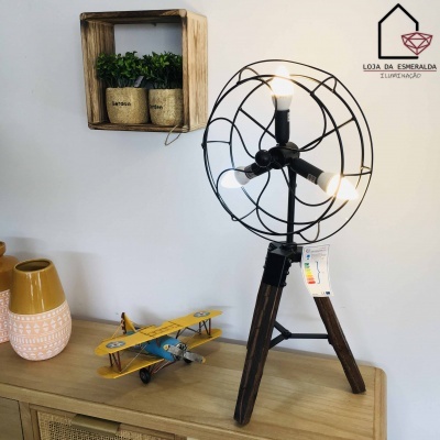 Abajur com tripé de madeira, estrutura metálica preta e três lâmpadas acesas sobre móvel de madeira com decoração ao fundo