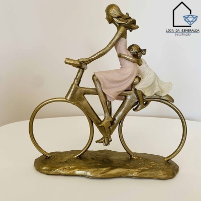Escultura de mulher e criança em bicicleta com acabamento dourado e vestidos pastel sobre superfície branca.