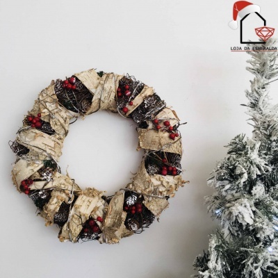 Coroa de Natal rústica com bagas vermelhas e casca de árvore pendurada numa parede ao lado de um pinheiro nevado.