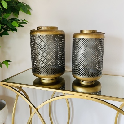 Duas lanternas decorativas douradas em mesa espelhada com planta ao lado