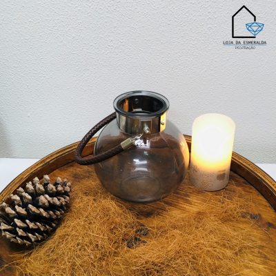 Lanterna de vidro com aro metálico e alça castanha junto a vela acesa e pinheiro seco numa bandeja de madeira