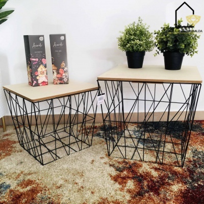 Mesas de centro com estrutura metálica preta geométrica e tampo de madeira clara, sobre tapete colorido, com plantas em vasos pretos e embalagens decorativas ao fundo