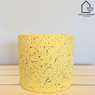 Vaso amarelo com manchas pretas e brancas sobre superfície branca com logotipo de loja na parede