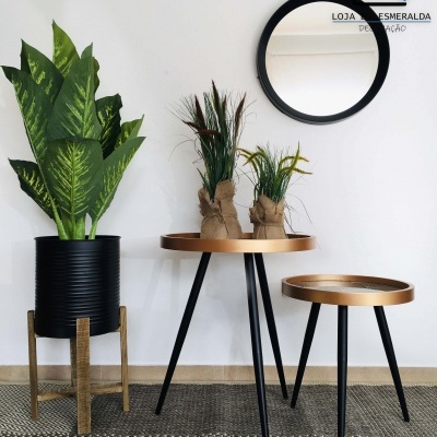 Mesa pequena redonda dourada com pernas pretas, cachepô preto com planta verde e espelho redondo na parede
