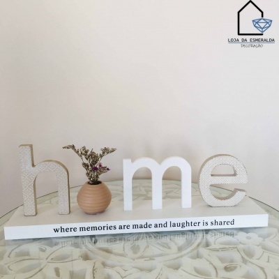 Peça decorativa em madeira com a palavra home e vaso com flores secas numa base branca com texto em inglês