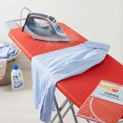 Tabuleiro de engomar vermelho com padrão de pontos brancos, ferro a vapor cinza, camisa azul e embalagem de forra para tábua de engomar.