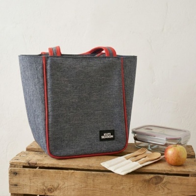 Sacola térmica cinza com detalhes vermelhos e etiqueta KUTI SEASON, talheres de madeira, marmita de vidro e maçã sobre madeira.