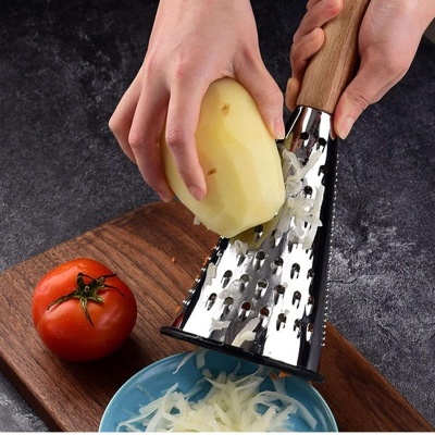 Ralador de metal com cabo de madeira a ralar batata sobre tigela azul, com tábua de madeira e tomate ao lado