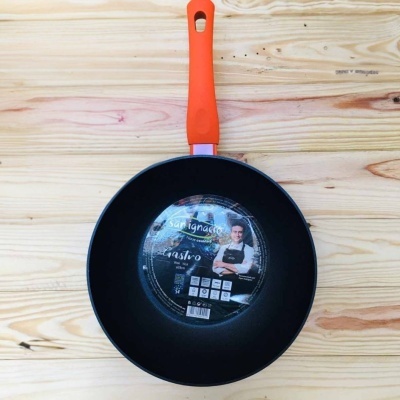 Wok preto com cabo laranja e etiqueta redonda em superfície de madeira.