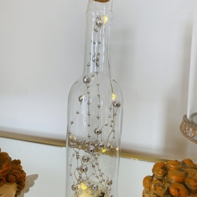 Garrafa de vidro com luzes LED e decoração prateada entre figuras angelicais