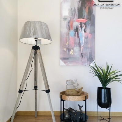 Abajur de tripé, mesa com estátuas, planta em vaso e quadro abstrato numa parede clara com logotipo